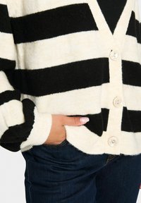Cardigan a righe nere e crema con scollo a V, dotato di cinque bottoni trasparenti e polsini a costine, abbinato a jeans in denim scuri.