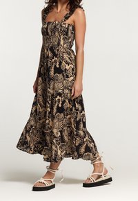 Floral-patterned maxi dress in black and beige with smocked bodice, square neckline, and ruffled straps; paired with strappy platform sandals.