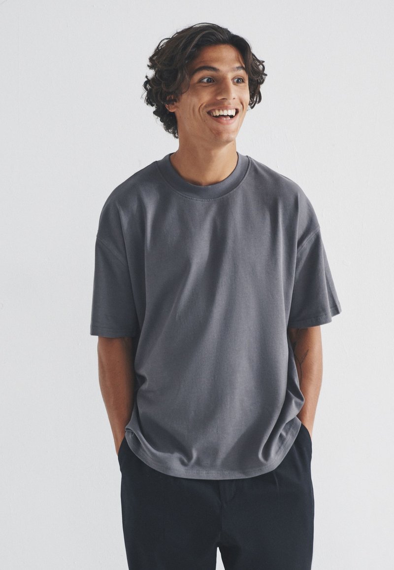 Jonge man met krullend donker haar die lacht, draagt een los grijs T-shirt en zwarte broek, staat met de handen in de zakken tegen een effen achtergrond.