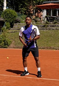 Tennis speler in een paarse en witte jersey, zwarte shorts en zwarte sneakers, met een tennisracket op een gravelbaan.