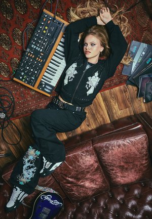 Black cropped jacket with graphic design, black pants with embroidered pattern, vintage synthesizer, brown leather couch, wooden floor.