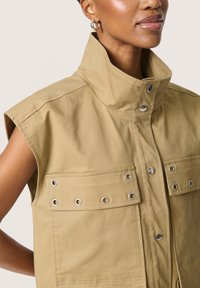 Beige mouwloos vest met een hoge kraag, twee voorzakken met oogje-details en drukknoopsluitingen, gemaakt van een stevige stof.
