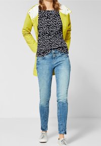 Žlutá voděodolná bunda, černý top s bílými puntíky, modré slim-fit džíny a bílé tenisky. Jednoduchý, ležérní outfit s vrstvenými barvami.