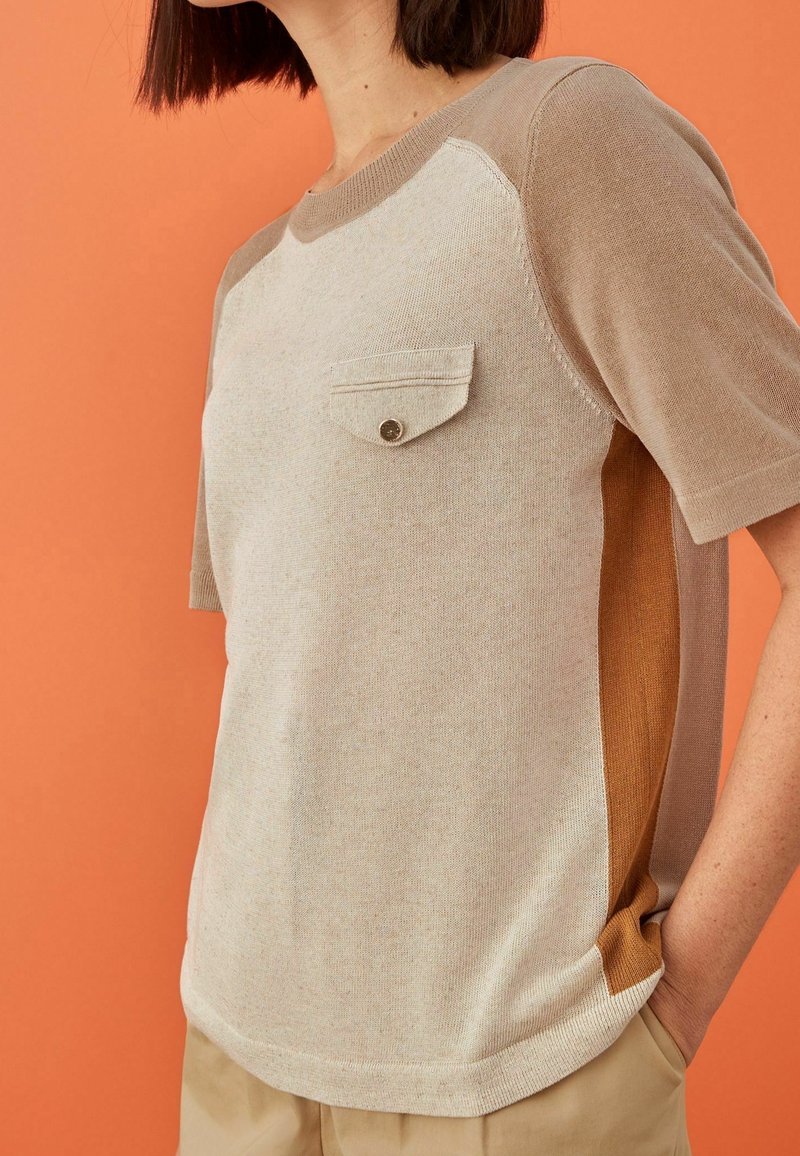 Femme portant un haut en maille beige et marron à manches courtes avec une petite poche poitrine boutonnée, debout devant un fond orange.