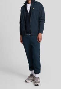 Mørkeblå zip-up jakke over en marineblå fleece, parret med matchende joggers og grå sneakers. Har lommer og en lille gul logo detalje.