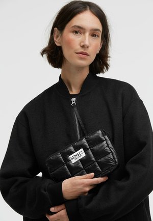 Schwarze gesteppte Clutch mit einem Steppmuster und einem weißen Etikett. Das Modell trägt eine schwarze Bomberjacke mit Reißverschluss.