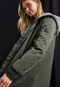 Pitkä oliivinvihreä takki, jossa on harmaa huppu, ribbikuvioiset hihansuut, vetoketjusuljenta ja patch vasemmassa hihassa. Sujuva, kevyt materiaali.