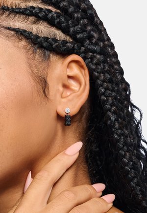 Steckerohrring in Form eines schwarzen Bären mit klarem Strassstein-Akzent, getragen am Ohrläppchen. Lockiges, strukturiertes Haar ist teilweise sichtbar.