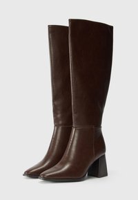 Bottines en cuir marron, montant jusqu'au genou, avec un bout pointu, un talon carré et une texture lisse ; dotées d'un design épuré et minimaliste.