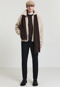 Giacca in shearling beige con colletto ampio, sciarpa lavorata a maglia marrone scuro, maglione scuro, pantaloni neri e stivaletti neri alla caviglia. Il look è completato da un berretto piatto.