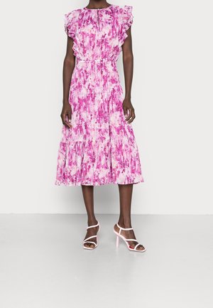 Femme portant une robe midi fleurie rose et blanche avec des manches à volants et des sandales à talons hauts roses à brides, debout sur un fond uni.