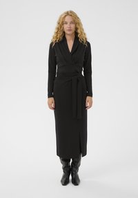 Juodas apvalios suknelė su giliu V formos kaklu, ilgomis rankovėmis, rišamu juosmens akcentu ir priekiu su plyšiu. Derinama su blizgausi juodais iki kelių batais.