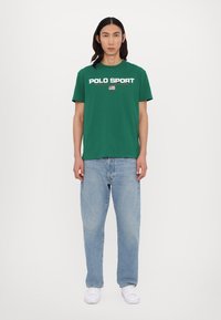 Camiseta de algodón verde de manga corta, con el logo "POLO SPORT" en blanco y rojo, combinada con jeans rectos de color azul claro y zapatillas blancas.