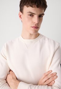 Lichtbeige sweatshirt met een gestructureerde afwerking, klassieke ronde halslijn en een kleine diagonale naaddetail bij de kraag. Slanke pasvorm.