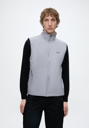Jonge man die een lichtgrijs mouwloos vest met een hoge kraag en een zwart shirt met lange mouwen draagt, staande tegen een eenvoudige witte achtergrond.