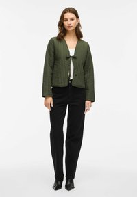 Vrouw in een olijfgroene gewatteerde jas met een strik aan de voorkant, witte top, zwarte high-waisted broek en zwarte puntige schoenen, staande tegen een witte achtergrond.