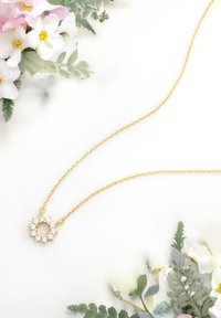 Guldkæde med en cirkulær vedhæng, der har ni klare rhinsten arrangeret i et blomsterdesign, sat mod en lys baggrund med blomster.