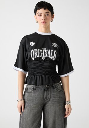 Zwart cropped T-shirt met witte accenten en ingesnoerde taille. Voorzien van bedrukte tekst en patches. Draagt losse grijze spijkerbroek en zilveren armband.