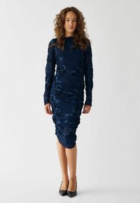 Rochie ajustată de culoare albastru închis, cu mâneci lungi, având broderie florală texturată și un design cu material strâns, care ajunge ușor sub genunchi.