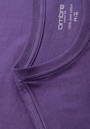 Lila Baumwoll-T-Shirt mit Rundhalsausschnitt und sichtbaren Nähdetails. Das Etikett trägt die Aufschrift "100 % reine Baumwolle" und hat eine weiche, glatte Textur.