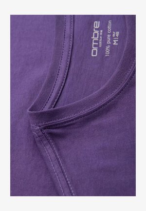 Lila Baumwoll-T-Shirt mit Rundhalsausschnitt und sichtbaren Nähdetails. Das Etikett trägt die Aufschrift "100 % reine Baumwolle" und hat eine weiche, glatte Textur.