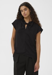 Giovane donna con capelli ricci che indossa un top nero a maniche corte con scollo annodato e pantaloni larghi neri, in piedi su uno sfondo bianco.