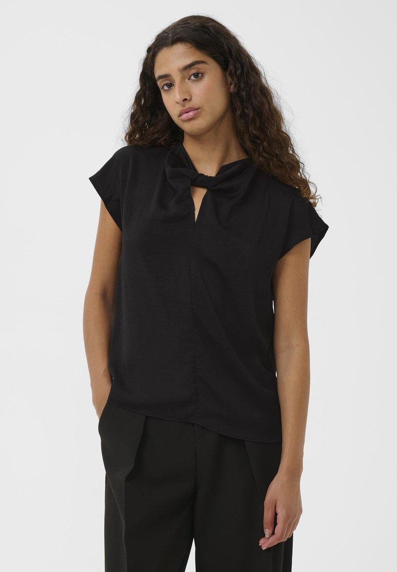 Giovane donna con capelli ricci che indossa un top nero a maniche corte con scollo annodato e pantaloni larghi neri, in piedi su uno sfondo bianco.