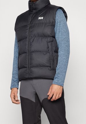 Mand iført sort dunvest med hvidt "HH"-logo over blå langærmet skjorte og to-tonet grå og sorte bukser.