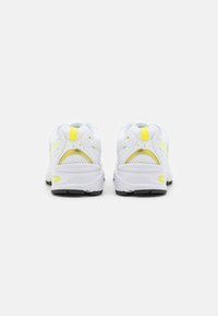 Vita träningsskor i meshmaterial, med gula accenter, rundad häl och texturerad sula. Bakifrån visas design- och färgdetaljer.