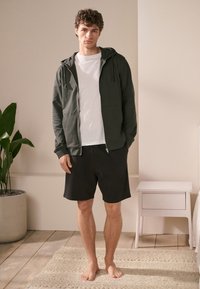 Donkergroene rits hoodie met een voorke pocket, gedragen over een wit T-shirt en zwarte shorts. Model staat op een licht houten vloer.