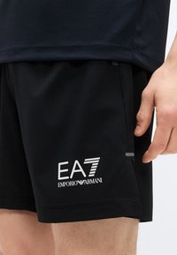 Μαύρο αθλητικό σορτς από αναπνεύσιμο ύφασμα, με λευκό λογότυπο EA7 και λείο υφή. Πλαϊνές τσέπες και μινιμαλιστικός σχεδιασμός.