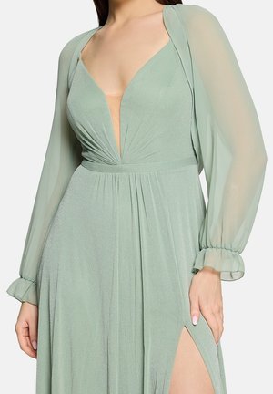 Femme portant une robe vert clair avec des manches longues transparentes, un décolleté en V profond, une taille plissée et une fente haute jusqu'à la cuisse.