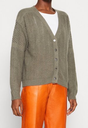 Personne à la peau foncée portant un cardigan en maille vert olive avec boutons par-dessus un haut blanc et un pantalon en cuir orange vif.