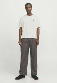 T-shirt jaune clair à manches courtes avec un petit graphisme noir, associé à un pantalon plissé gris foncé et des chaussures noires.