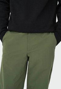 Schwarzes Langarmshirt, kombiniert mit olivgrünen Hosen. Die Hosen zeichnen sich durch eine glatte Textur und Nähte aus, mit Taschen an den Seiten.