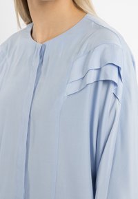 Blusa azzurro chiaro con chiusura a bottoni e dettagli a strati con volant sulle maniche. Collo e spalla del modello parzialmente visibili.