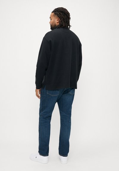 Μαύρο φούτερ με άνετη εφαρμογή, μακριά μανίκια και πλαϊνές χαρακιές· συνδυασμένο με μπλε τζιν τζιν και λευκά αθλητικά παπούτσια.