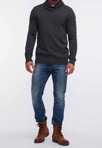 Donkergrijze gebreide trui met een sjaalkraag, gecombineerd met blauwe denim jeans en bruine veterlaarzen. Heeft geribde manchetten en zoom.