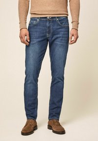 Hombre con jeans ajustados azules, suéter beige y zapatos de ante marrón con cordones frente a un fondo beige liso.