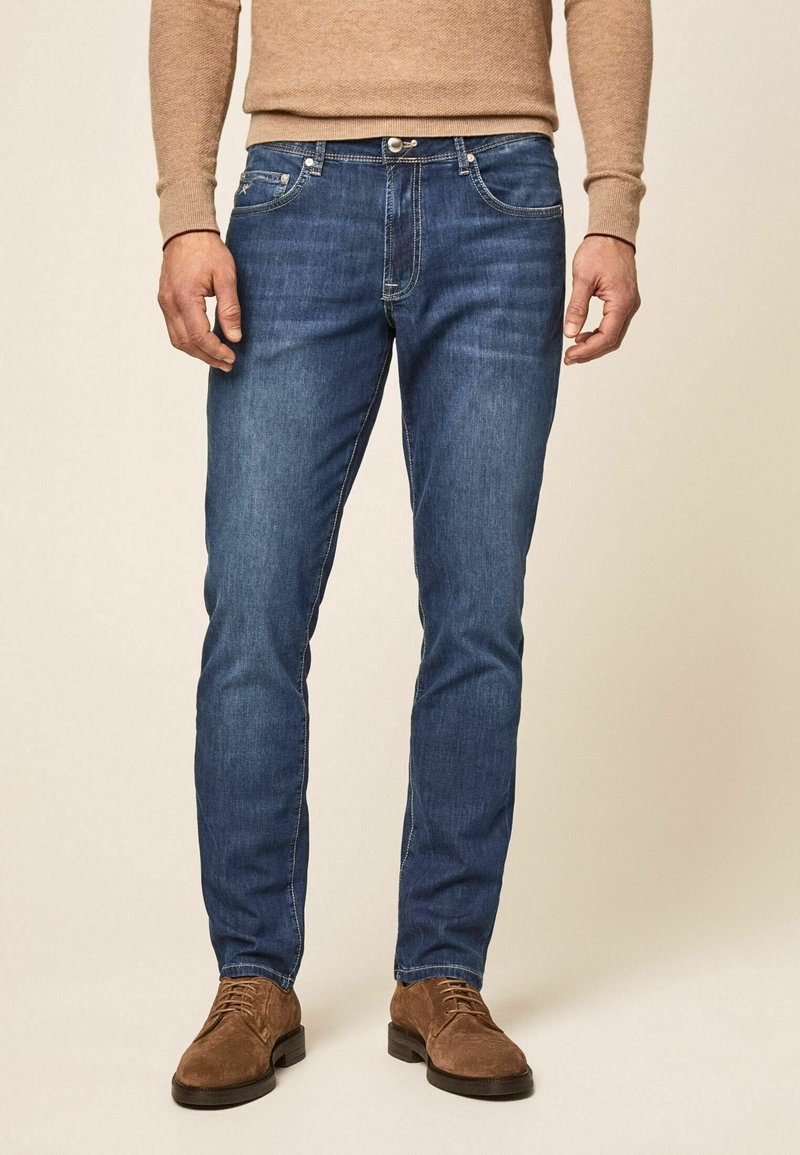 Hombre con jeans ajustados azules, suéter beige y zapatos de ante marrón con cordones frente a un fondo beige liso.