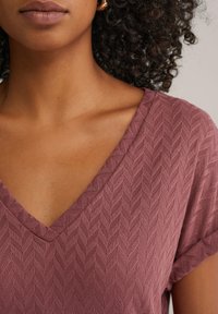 Haut rose avec un décolleté en V, présentant un motif en chevrons texturé et des manches courtes. Le tissu semble léger et respirant.