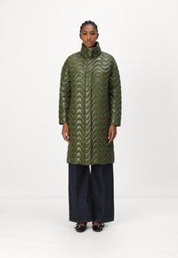 Padded olivgrüner Mantel mit welligem, gestepptem Muster, hohem Kragen und langen Ärmeln, kombiniert mit weit geschnittenen, dunklen Jeans und schwarzen Schuhen.