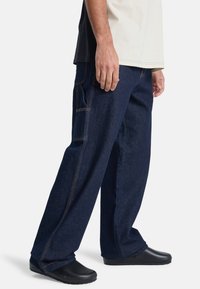 Homme portant un jean en denim bleu foncé ample avec une poche latérale et des chaussures noires sans lacets, debout devant un fond blanc.