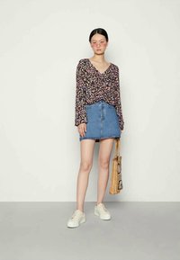 Top de mangas longas preto com padrão floral, saia de ganga e sapatilhas brancas. A modelo segura uma bolsa tecida com acentos geométricos. Fundo cinza.