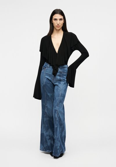 Γυναίκα που στέκεται φορώντας μαύρο τοπ με βαθύ V και βολάν, με μακριά φαρδιά μανίκια και ψηλόμεσο φαρδύ τζιν μπλε χρώματος, μπροστά σε απλό φόντο.