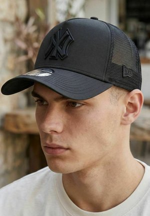 Young man wearing a black mesh baseball cap with a raised NY logo on front, facing slightly left, indoors with blurred background.