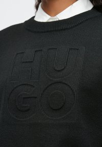 Κοντινό πλάνο ενός μαύρου πλεκτού πουλόβερ πάνω από ένα λευκό πουκαμισάκι, με διακριτό ανάγλυφο κείμενο "HUGO" στο στήθος.