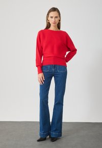 Pull rouge côtelé à manches longues, associé à un jean en denim évasé. Des escarpins noirs à bout pointu complètent la tenue. Fond simple.