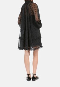 Schwarzes, durchsichtiges Kleid mit Punkten, langen Puffärmeln, Rüschendetails und gerafftem Ausschnitt. Getragen mit schwarzen Lacklederschuhen.