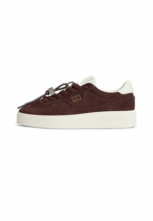 Zapatilla baja de ante marrón oscuro con suela blanca, detalle blanco en el talón, cordones con patrón y logo sutil en el lateral y la suela.